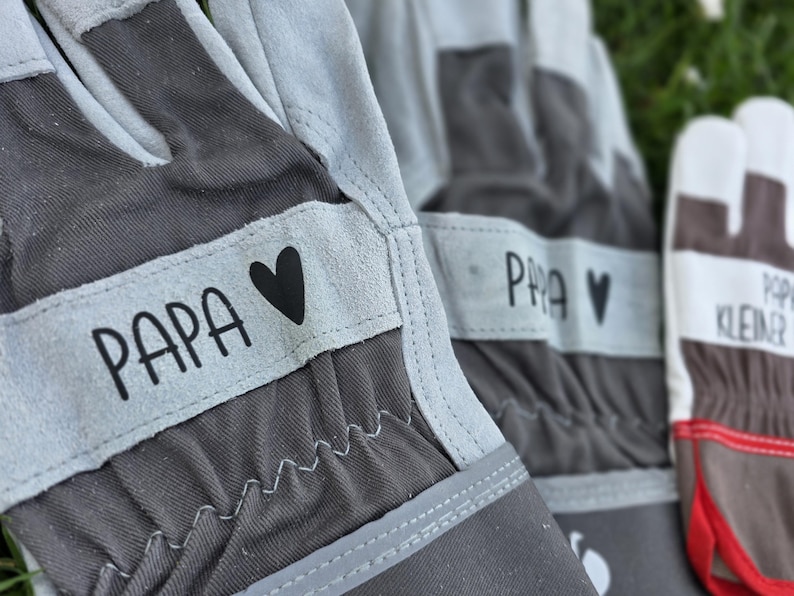 May include: Close-up of gray and light gray gardening gloves. The gloves have the word "PAPA" printed on a light gray band, along with a black heart. The gloves are made of a durable material and are designed for gardening.