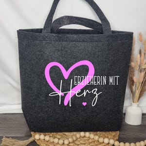 Könnte beinhalten: Eine graue Filztasche mit einem pinkfarbenen Herzen und dem Text "Erzieherin mit Herz" in weiß.