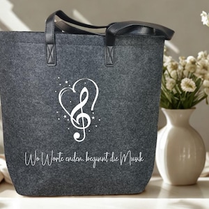 Könnte beinhalten: Graue Filz-Tragetasche mit schwarzen Griffen und einem weißen Noten-Design. Das Design umfasst einen Violinschlüssel in Herzform, umgeben von Sternen. Der Text "Wo Worte enden, beginnt die Musik" ist unten aufgedruckt.