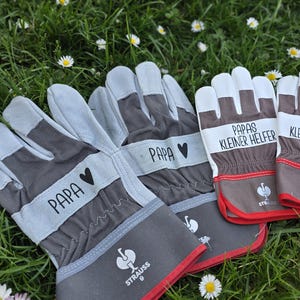 May include: Gray and brown work gloves with the word "PAPA" and a heart symbol printed on the wrist. Two pairs of gloves have the text "PAPAS KLEINER HELFER" printed on them. The gloves have red trim and are on a bed of grass.