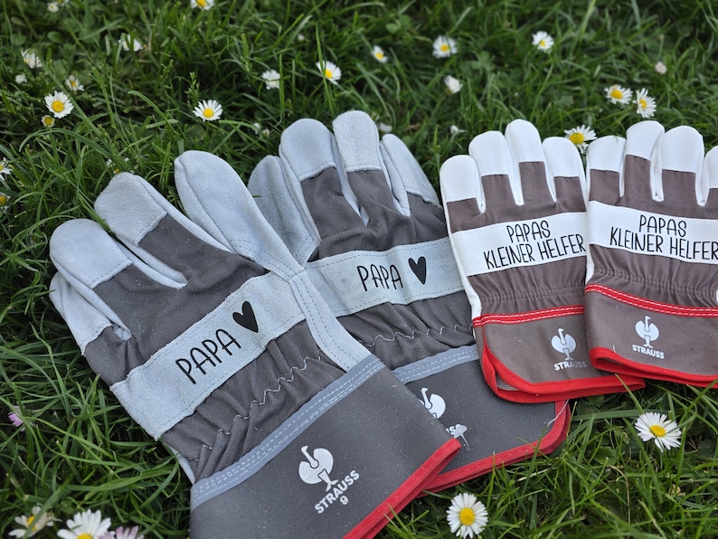 May include: Gray and brown work gloves with the word "PAPA" and a heart symbol printed on the wrist. Two pairs of gloves have the text "PAPAS KLEINER HELFER" printed on them. The gloves have red trim and are on a bed of grass.
