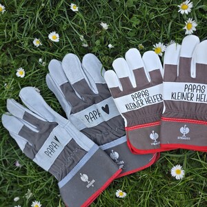May include: A set of work gloves with gray, white, and brown accents, resting on a bed of green grass and small white flowers. The gloves have the word "PAPA" and "Papas kleiner Helfer" printed on them.