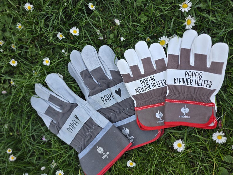 May include: A set of work gloves with gray, white, and brown accents, resting on a bed of green grass and small white flowers. The gloves have the word "PAPA" and "Papas kleiner Helfer" printed on them.