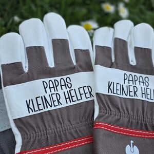 May include: A pair of work gloves with white fingertips and a brown and white design. The gloves have the text "PAPAS KLEINER HELFER" printed on them in black. Red stitching accents the bottom of the gloves.