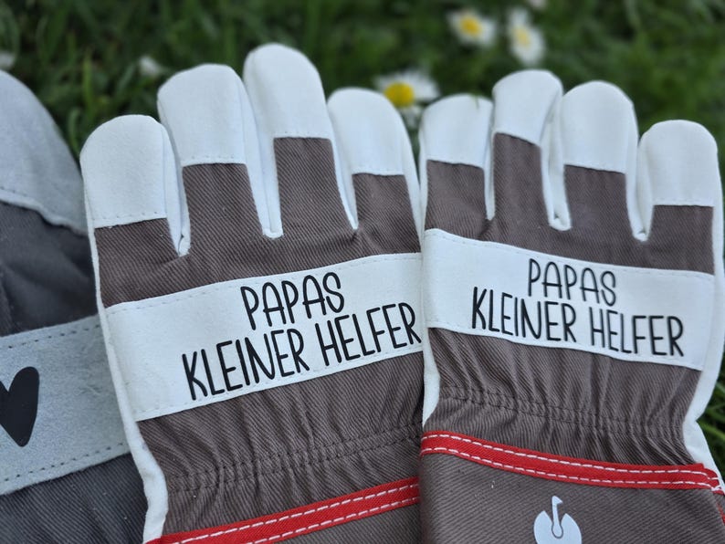 May include: A pair of work gloves with white fingertips and a brown and white design. The gloves have the text "PAPAS KLEINER HELFER" printed on them in black. Red stitching accents the bottom of the gloves.
