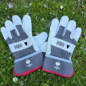 May include: A pair of work gloves with the word "PAPA" and a black heart printed on the gray band. The gloves are light blue and gray with a red trim. The gloves are lying on a bed of green grass.