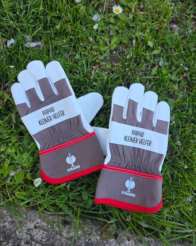 May include: A pair of work gloves with white palms and fingers, brown backs, and red trim. The text "PAPAS KLEINER HELFER" is printed on a white band across the back of each glove. The gloves are resting on a bed of green grass.