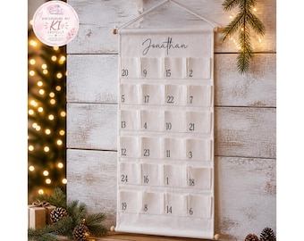 May include: A vertical off-white fabric advent calendar hanging from a wooden rod with a string. The calendar features twenty-four individual pockets, each printed with a black number from one to twenty-four, and the name Jonathan at the top.
