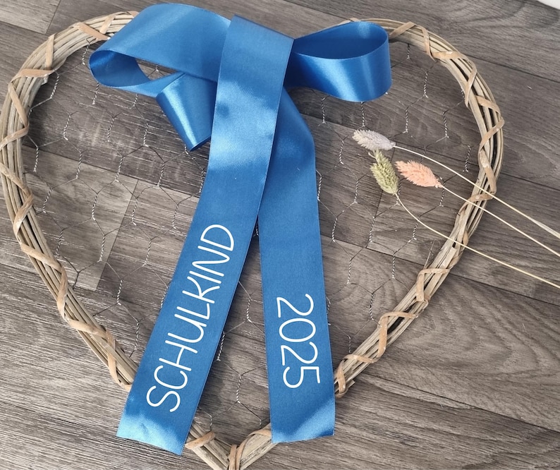 May include: A blue satin ribbon tied in a bow, featuring the white text SCHULKIND and 2025 printed on the hanging tails. The ribbon is positioned over a heart-shaped frame made of woven light brown wicker and wire mesh with dried grass stems.