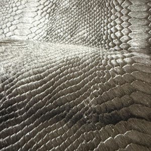 Op de afbeelding: Close-up van een zwarte, getextureerde stof met een patroon dat lijkt op reptielenschubben. Het materiaal heeft een subtiele glans, met variaties in licht en schaduw over het oppervlak. De textuur lijkt verhoogd en driedimensionaal.