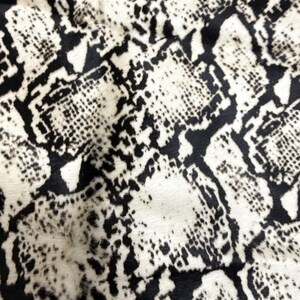Könnte beinhalten: Nahaufnahme eines Textils mit Schlangenmuster. Das Design zeigt ein sich wiederholendes Muster aus schwarzen und weißen Schuppen, wodurch ein strukturiertes Aussehen entsteht. Der Stoff hat eine natürliche, organische Haptik.