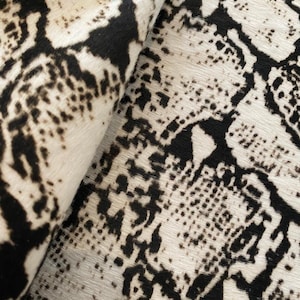 Könnte beinhalten: Nahaufnahme eines Teppichs mit Schlangenmuster. Der Teppich hat einen weißen Hintergrund mit schwarzen Markierungen, wodurch ein strukturiertes Aussehen entsteht. Der Teppich ist teilweise gerollt und zeigt die Unterseite.