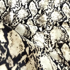 Könnte beinhalten: Nahaufnahme eines Stoffes mit Schlangenmuster. Der Stoff weist ein sich wiederholendes Design aus schwarzen und weißen Schuppen auf, wodurch ein strukturiertes Aussehen entsteht. Das Material ist gefaltet, was die Kontinuität des Musters und die Flexibilität des Stoffes zeigt.