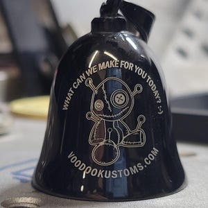 May include: Black bell-shaped ornament with a voodoo doll design and the text "WHAT CAN WE MAKE FOR YOU TODAY? :) " and "VOODOOKUSTOMS.COM" in silver. The ornament has a loop at the top.