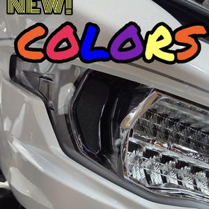 May include: Close-up of a white motorcycle with the text "NEW! COLORS" in colorful bubble letters. The motorcycle has a shiny, reflective surface and a black accent around the headlight.