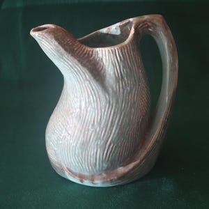 May include: A ceramic pitcher with a textured, wood-grain-like surface. The pitcher is primarily a muted gray-green color with brown accents at the base and rim. It has a curved handle and a spout for pouring.