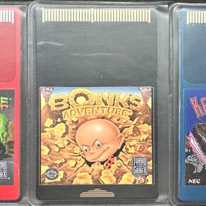 Puede incluir: Tres cartuchos de videojuegos HuCard TurboGrafx-16 vintage. Los cartuchos están en estuches de plástico transparente. Los juegos son "Splatterhouse" en un cartucho rojo, "Bonk's Adventure" en un cartucho negro y "Keith Courage in Alpha Zones" en un cartucho azul.