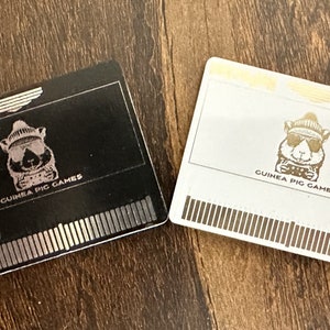 Puede incluir: Dos cartuchos de videojuegos, uno negro y otro blanco, con un conejillo de indias con gafas de sol y gorro. El texto "GUINEA PIG GAMES" está debajo. La palabra "HUSTUB" está en el cartucho negro.