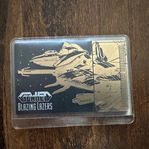 May include: A gold and black card with a spaceship design and the text "TUNNEL BLAZING LAZERS". The card is in a clear plastic sleeve. The background depicts a starry night sky.