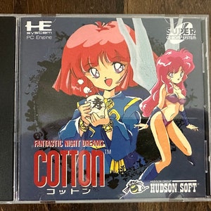Puede incluir: Caja de videojuego para PC Engine de "Cotton: Fantastic Night Dreams". La carátula presenta a dos mujeres de estilo anime con cabello rojo, sobre un fondo azul oscuro. El título del juego se muestra en rojo, con texto japonés debajo. La caja está en un estuche de plástico transparente.