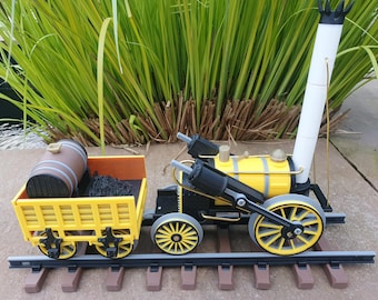 May include: A toy train set featuring a yellow and black steam engine with a white smokestack and a yellow coal car. The train is on a brown track, with a brown barrel in the coal car. The train is set against a backdrop of green foliage.