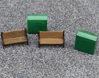 May include: Three miniature model buildings, two are green and one is brown. The green buildings are small sheds with a pitched roof. The brown building is a bench with black metal legs.
