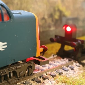 May include: A model railway scene with a teal train carriage, a white logo, and an orange stripe. A red signal light is lit in the background. The train is on a track with a gravel bed.