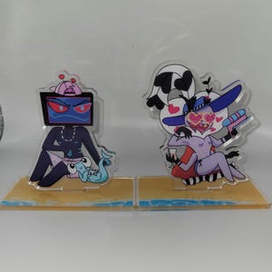 Może przedstawiać: Dwie akrylowe figurki przedstawiające postacie z kreskówek. Jedna ma głowę telewizora i nosi różowy kapelusz i kąpielówki. Druga ma twarz w kształcie serca, duży kapelusz i trzyma pistolet na wodę. Obie stoją na podstawie o tematyce plażowej.