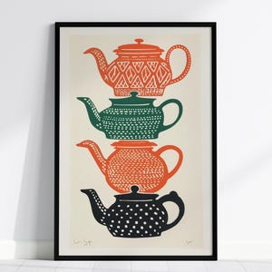 Op de afbeelding: Een ingelijste verticale kunstdruk met een stapel van vier gestileerde theepotten in een kolom. De bovenste theepot is oranje met een ruitpatroon, de tweede is groen met stippen, de derde is oranje met een textuurpatroon en de onderste is zwart met witte stippen.