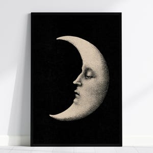 May include: A framed wall art print featuring a cream-colored crescent moon with a human face in profile, set against a solid black background. The moon illustration uses stippling and fine line shading to define the facial features and texture.