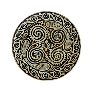 Puede incluir: Un plato redondo de cerámica con fondo negro y un diseño geométrico blanco y marrón. El diseño presenta patrones en espiral y puntos.