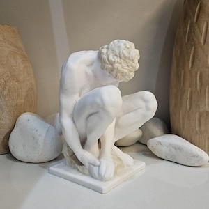 May include: White marble sculpture of a nude male figure in a seated pose. The figure is leaning forward with its head resting on its hand. The sculpture is on a white marble base.