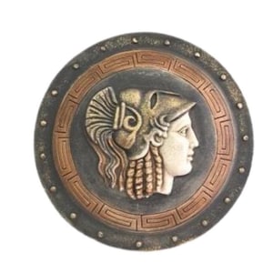 Puede incluir: Un escudo redondo decorativo con un acabado en bronce y cobre. El escudo presenta una imagen en relieve detallada de la cabeza de una mujer con casco y una trenza suelta. El escudo está decorado con un patrón geométrico repetitivo alrededor del borde.