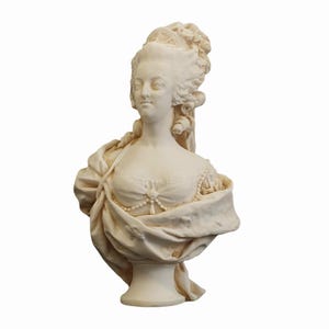 Op de afbeelding: Een crèmekleurige buste-sculptuur van een vrouw met een uitgebreid kapsel en gedrapeerde kleding. De sculptuur toont een gedetailleerde weergave van het gezicht van de vrouw, met een parelketting en een gedrapeerde sjaal.