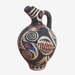 Puede incluir: Una jarra de cerámica negra con diseños pintados en blanco, rojo y azul. La jarra tiene un cuerpo redondeado y un pico corto y curvo.