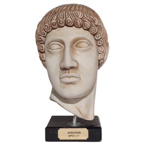 Könnte beinhalten: Cremefarbene Büste des Apollo mit detaillierten Gesichtszügen und lockigem braunem Haar. Die Skulptur ist auf einem schwarzen rechteckigen Sockel mit einem goldenen Etikett mit der Aufschrift "ΑΠΟΛΛΩΝ APOLLO" montiert.