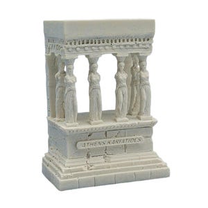 Puede incluir: Una réplica en miniatura blanca del Erecteión, un templo griego. La estructura presenta columnas y estatuas de mujeres, con el texto "ATHENS KARIATIDES" en la base. La maqueta arquitectónica está hecha de un material claro.