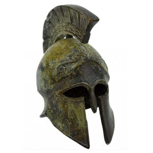 May include: A bronze-colored Spartan helmet with a crest and eye openings. The helmet has a weathered appearance with green and brown tones, and decorative patterns near the crest. The helmet is a historical replica.