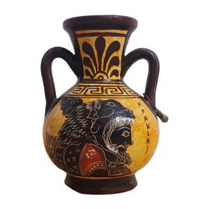 Puede incluir: Jarrón de cerámica pintado a mano, de estilo antiguo, con una combinación de colores amarillo y marrón. El jarrón presenta una ilustración negra de una figura con cabeza de león, con la palabra "PAKLIN" escrita en el lateral. Tiene dos asas y un borde marrón oscuro.