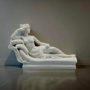 May include: A white marble sculpture of a woman reclining on a chaise longue. The sculpture is detailed with intricate carvings and a flowing drapery.