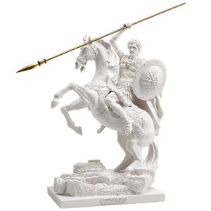 May include: A white statue of Alexander the Great riding a rearing horse on a rocky base. The figure wears armor and holds a round shield with a star emblem. A long, thin, gold-colored spear is held aloft. A plaque reads ALEXANDER THE GREAT.