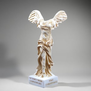 Puede incluir: Una estatua color crema de la Victoria de Samotracia. La estatua sin cabeza tiene alas extendidas y drapeado fluido. La base de la estatua tiene las palabras "VICTORY FROM SAMOTHRAKI" y "NIKH ΣΑΜΟΘΡΑΚΗΣ".