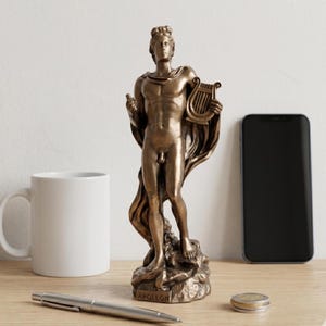 Può includere: Statua in bronzo di una figura maschile che tiene una lira e un piccolo oggetto. La statua è su una base rocciosa con l'iscrizione "APOLLON". Sono visibili anche una tazza bianca, una penna, uno smartphone e delle monete.