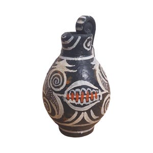Puede incluir: Jarrón de cerámica negro con diseños pintados en blanco y naranja. El jarrón tiene un asa curva única.