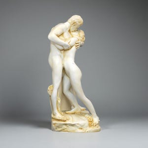 Può includere: Una scultura color crema raffigurante una coppia che si abbraccia e si bacia. Le figure nude hanno tratti del viso dettagliati e definizione muscolare. La scultura è montata su una base strutturata, suggerendo un tema classico.