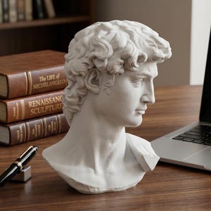 May include: A white plaster bust of Michelangelo's David, showcasing the sculpture's detailed facial features and curly hair. The bust is positioned on a wooden surface, near a laptop and books about Renaissance art.