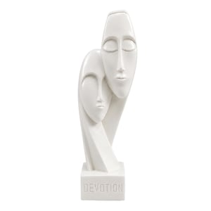 Puede incluir: Una escultura blanca lisa que presenta dos rostros estilizados y alargados colocados uno sobre otro. Los rostros tienen los ojos cerrados y expresiones neutras. La base es un bloque rectangular con la palabra DEVOTION grabada en relieve.