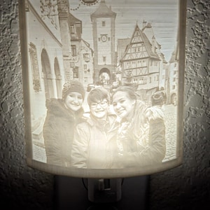 May include: A white ceramic night light with a 3D image of three people standing in front of a European city street. The image is lit from behind, creating a glowing effect. The text "Nurnberg" is visible in the image.