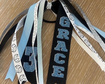 May include: Black and blue volleyball hair tie with the name "GRACE" and the number "3" on it. The hair tie has multiple ribbons with white volleyball designs.