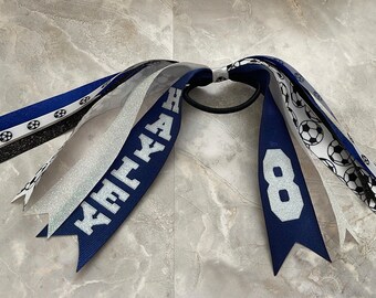 May include: A soccer-themed hair accessory featuring a black elastic band with multiple ribbons. The ribbons include navy blue, silver glitter, and black, with soccer ball patterns and the name "KAYLA" and the number "8" in white.